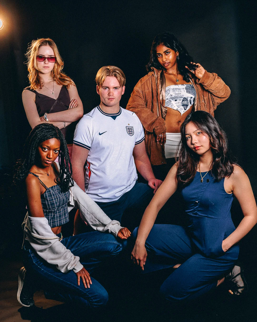 A diverse group of five young adults posing in stylish outfits against a dark studio background.