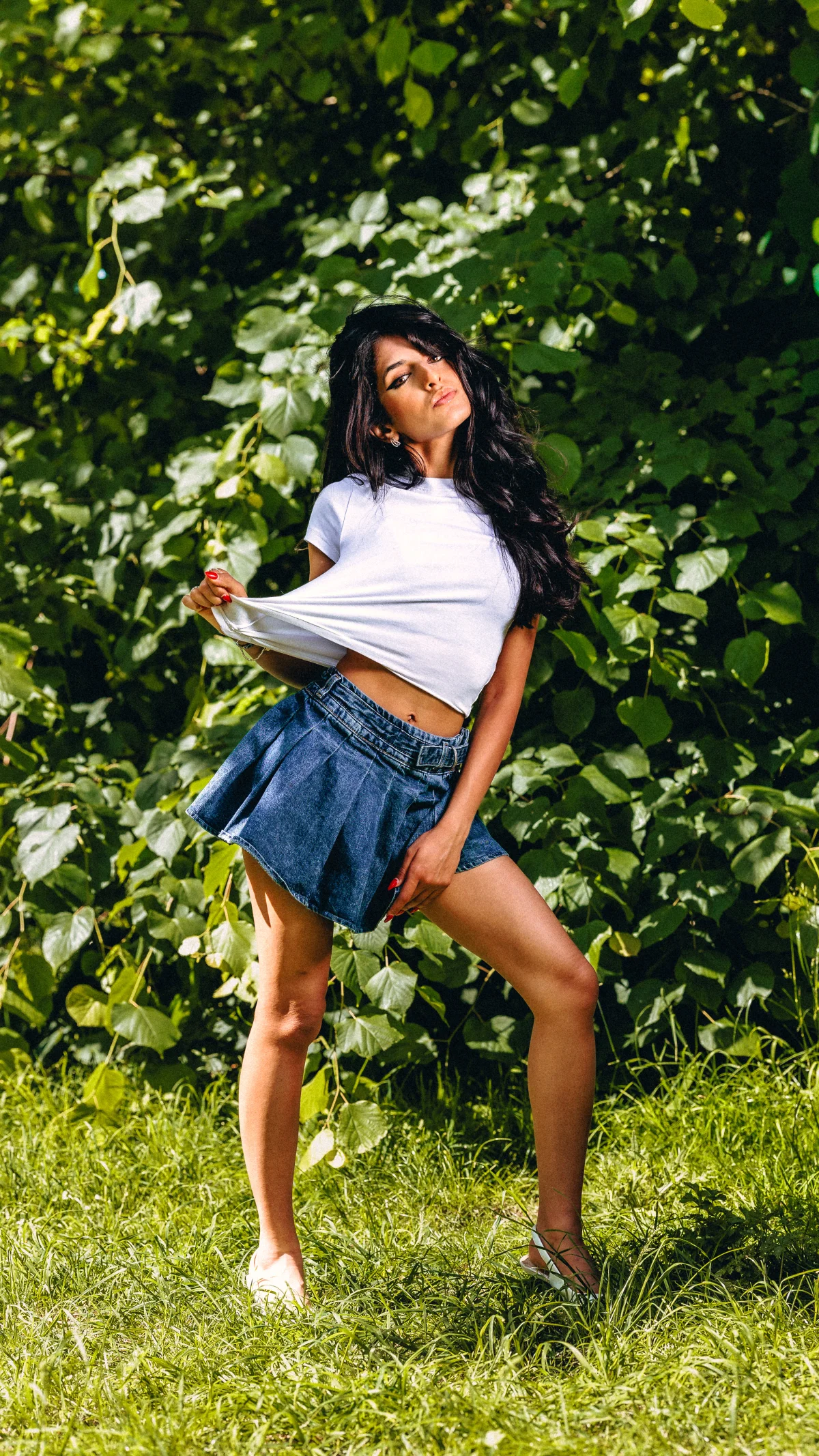 A woman with long dark hair posing in a white t-shirt and denim mini skirt against a lush green foliage background.