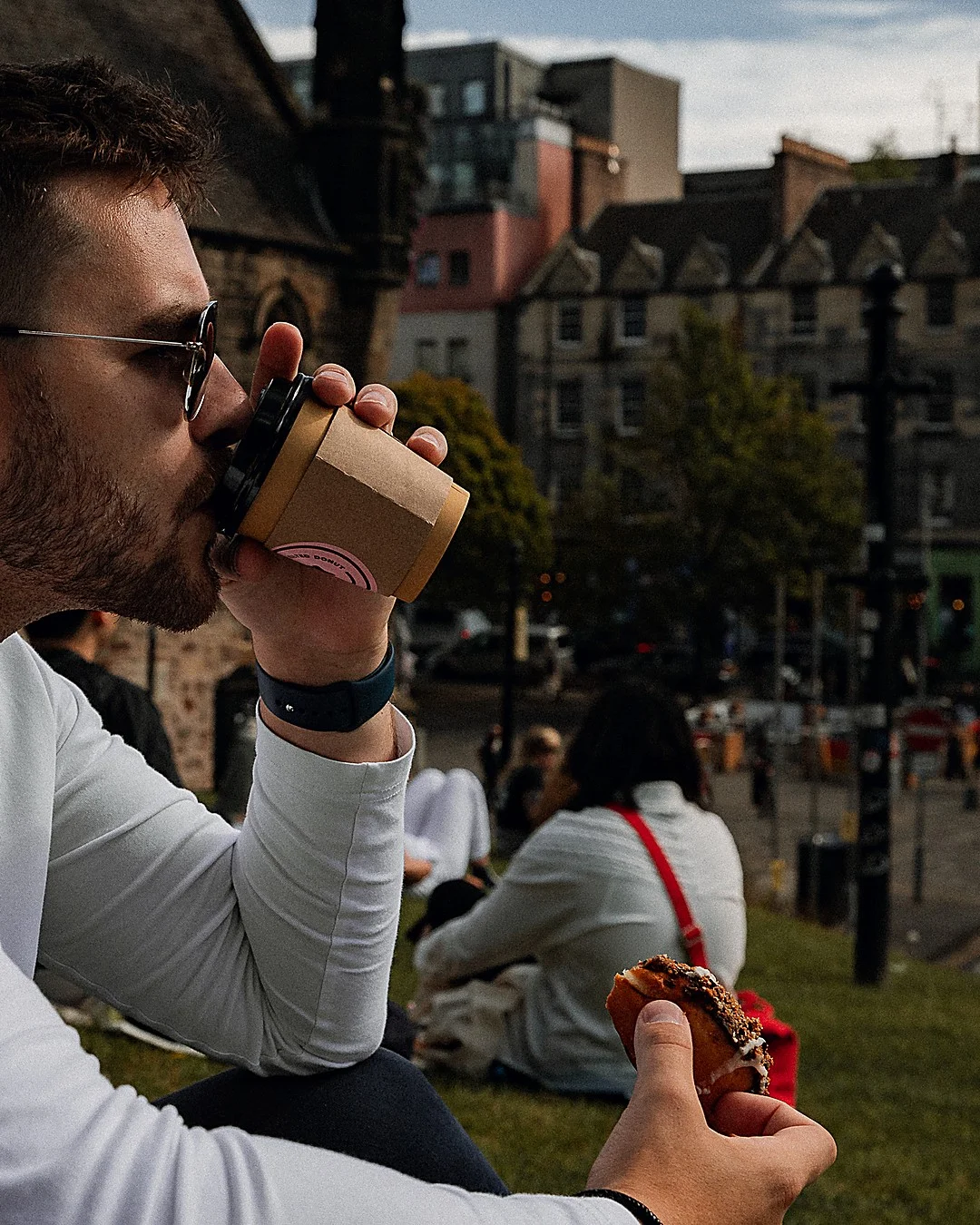 Side profile of a man in sunglasses and white shirt drinking coffee and holding a pastry outdoors.