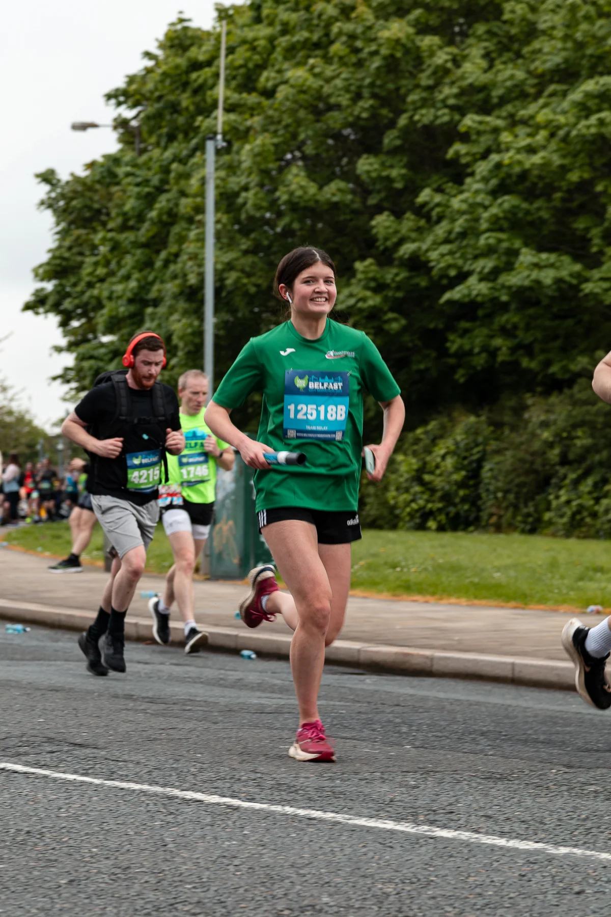 A woman in a green shirt runs during a race on a paved road with lush green trees in the background.