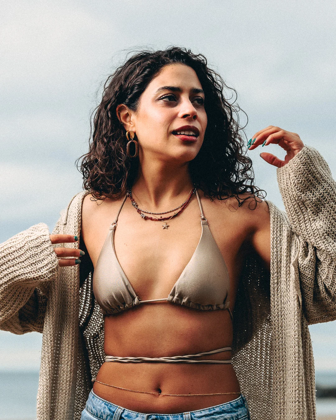A woman with curly dark hair in a beige bikini top and oversized cardigan posing outdoors.