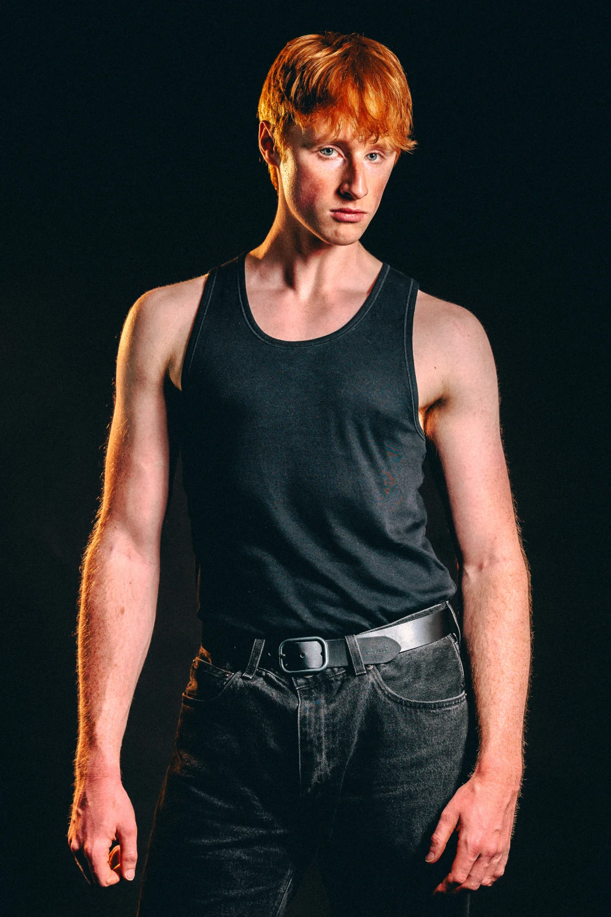 Young man with red hair in a black tank top and jeans against a dark background.