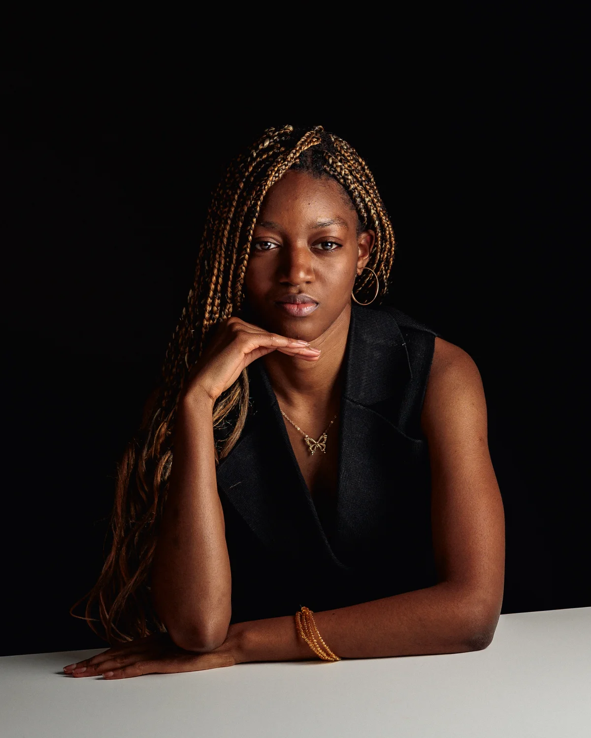 A woman with long blonde braids in a black sleeveless top posing against a dark background.