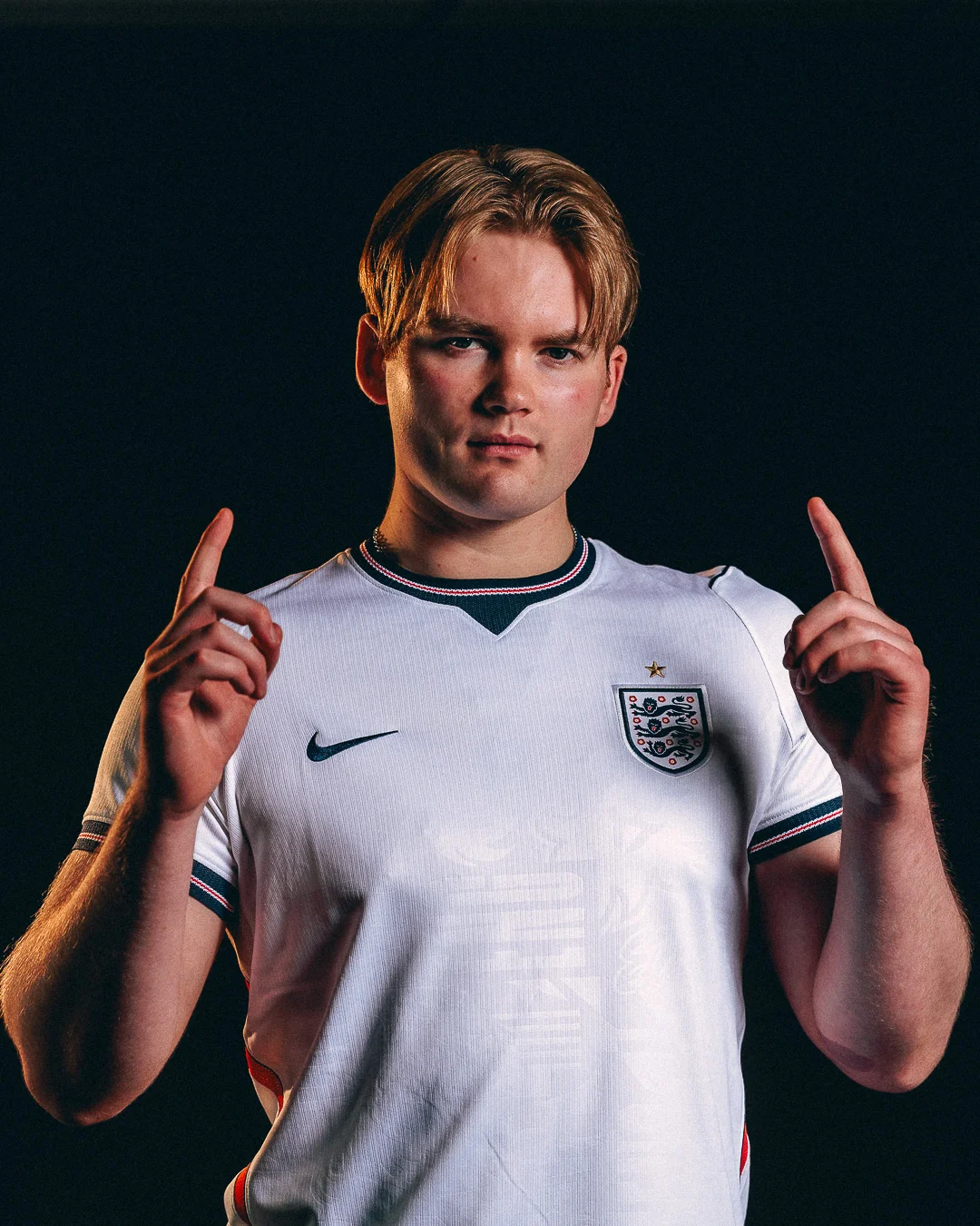 Young man in an England football jersey posing against a black background with fingers pointing up.