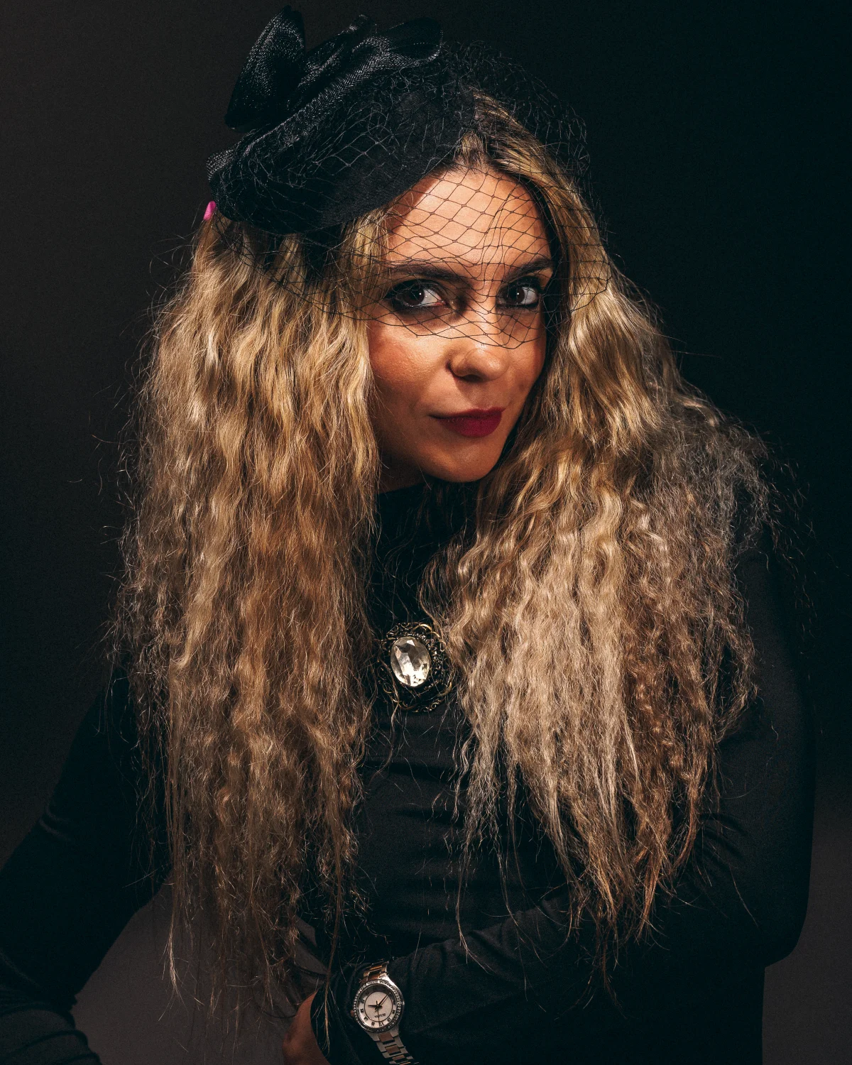 A woman with long curly blonde hair wearing a black veil hat and a black top with a large brooch.