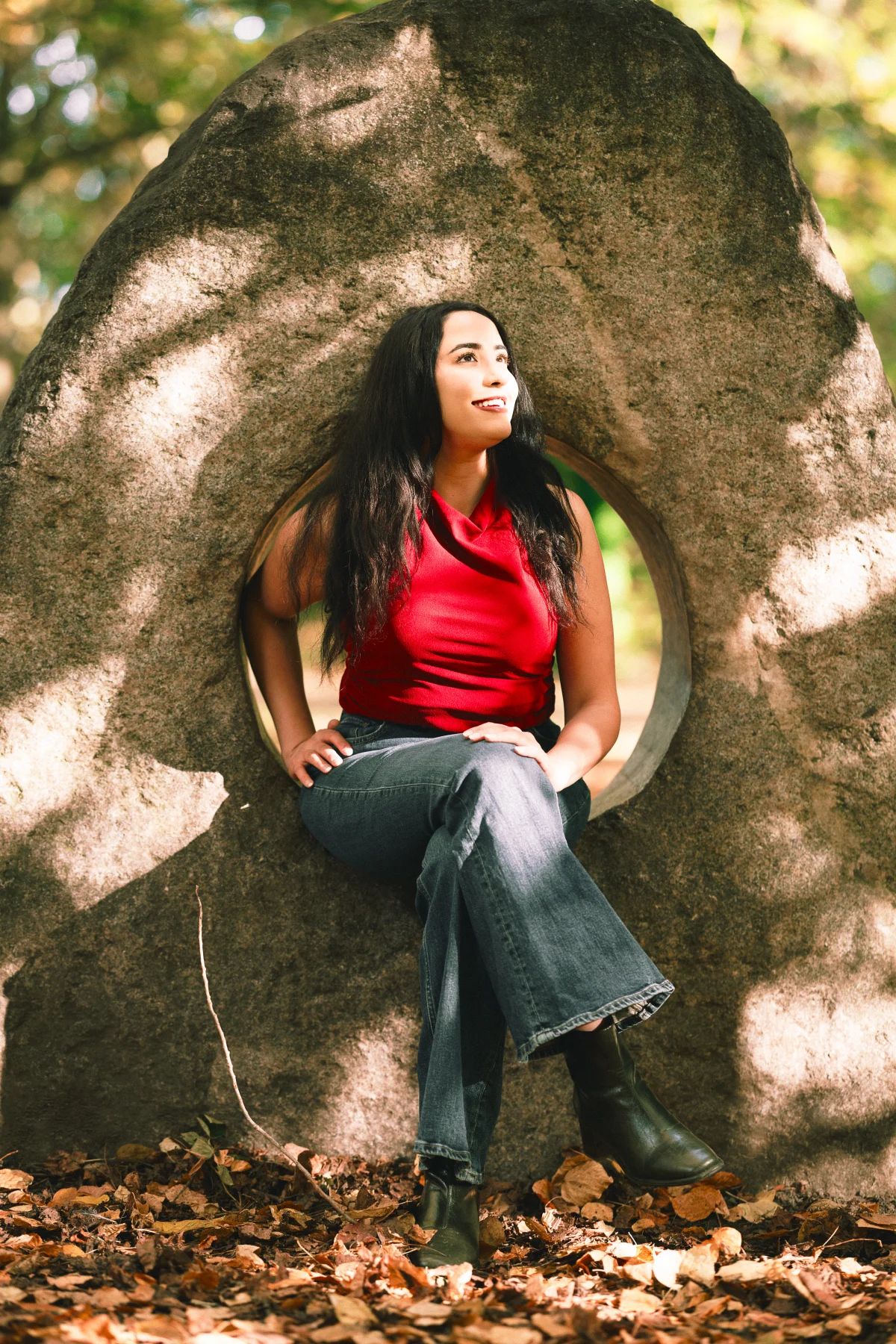 A woman in a red top and blue jeans sitting inside a circular opening of a large rock outdoors.