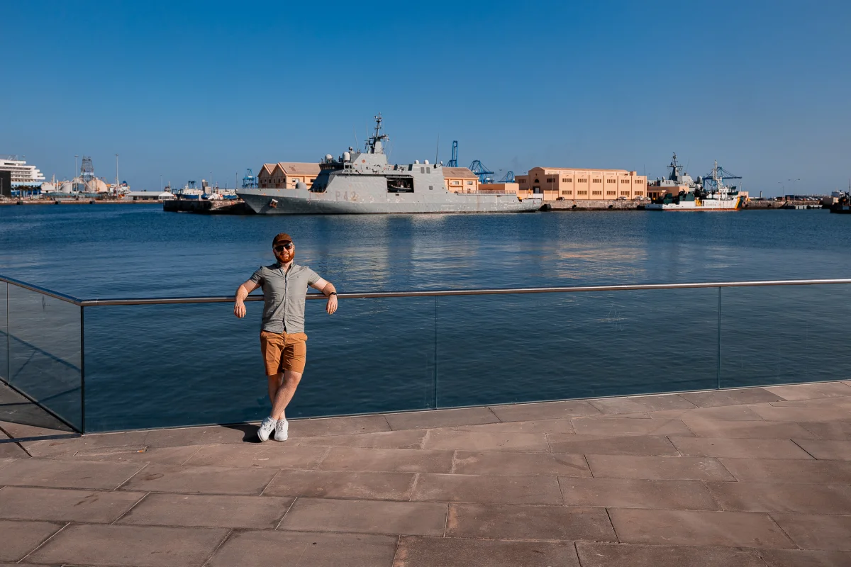 A man in a grey shirt and orange shorts leaning against a glass railing at a harbor with a large naval ship behind him.