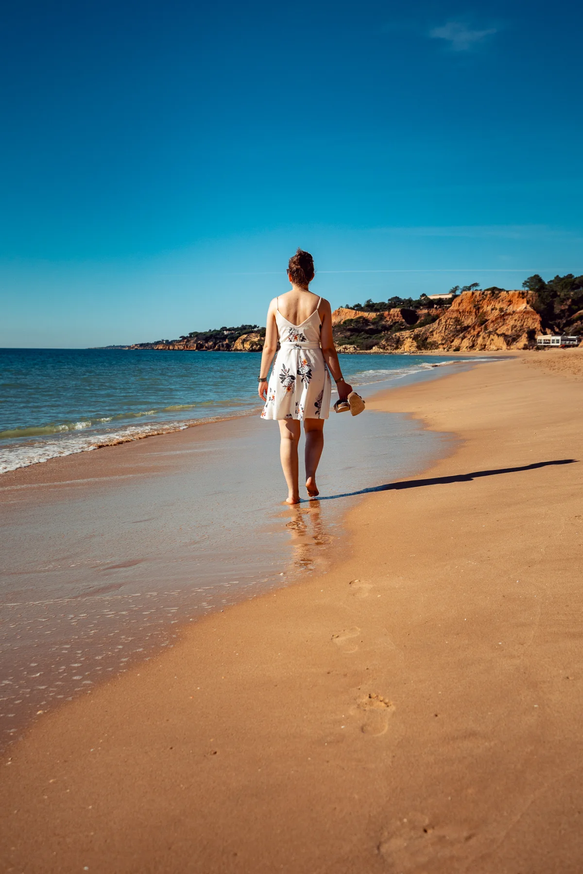 Woman in a white summer dress walking away along a sandy beach towards distant cliffs under a clear blue sky.
