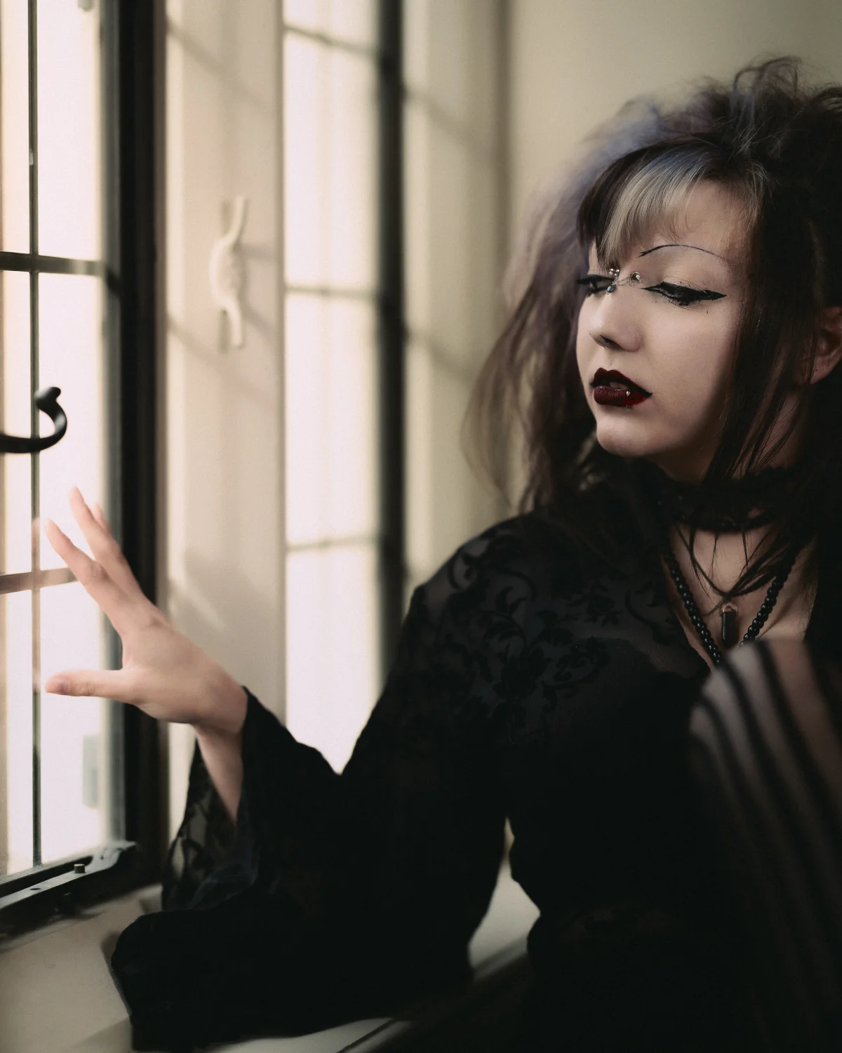 A woman with gothic makeup and black lace attire gazing thoughtfully by a window in moody lighting.