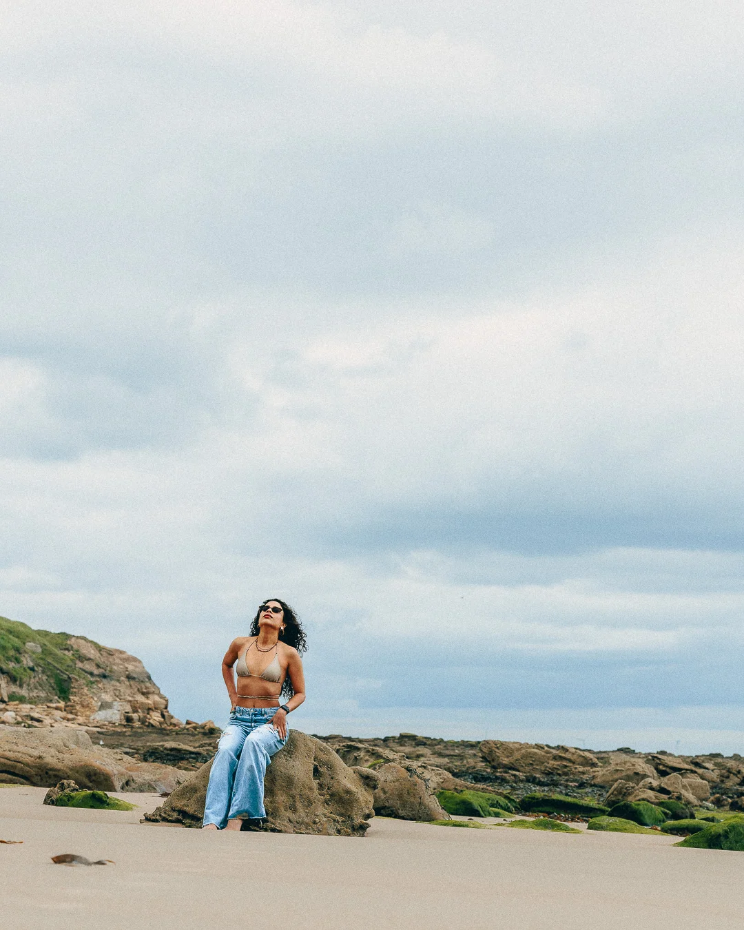 A woman in a gold bikini top and light blue jeans posing on coastal rocks under an overcast sky.