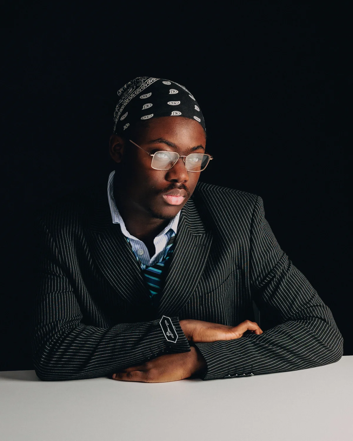 A man wearing glasses, a black bandana, and a pinstripe suit jacket against a solid black background.