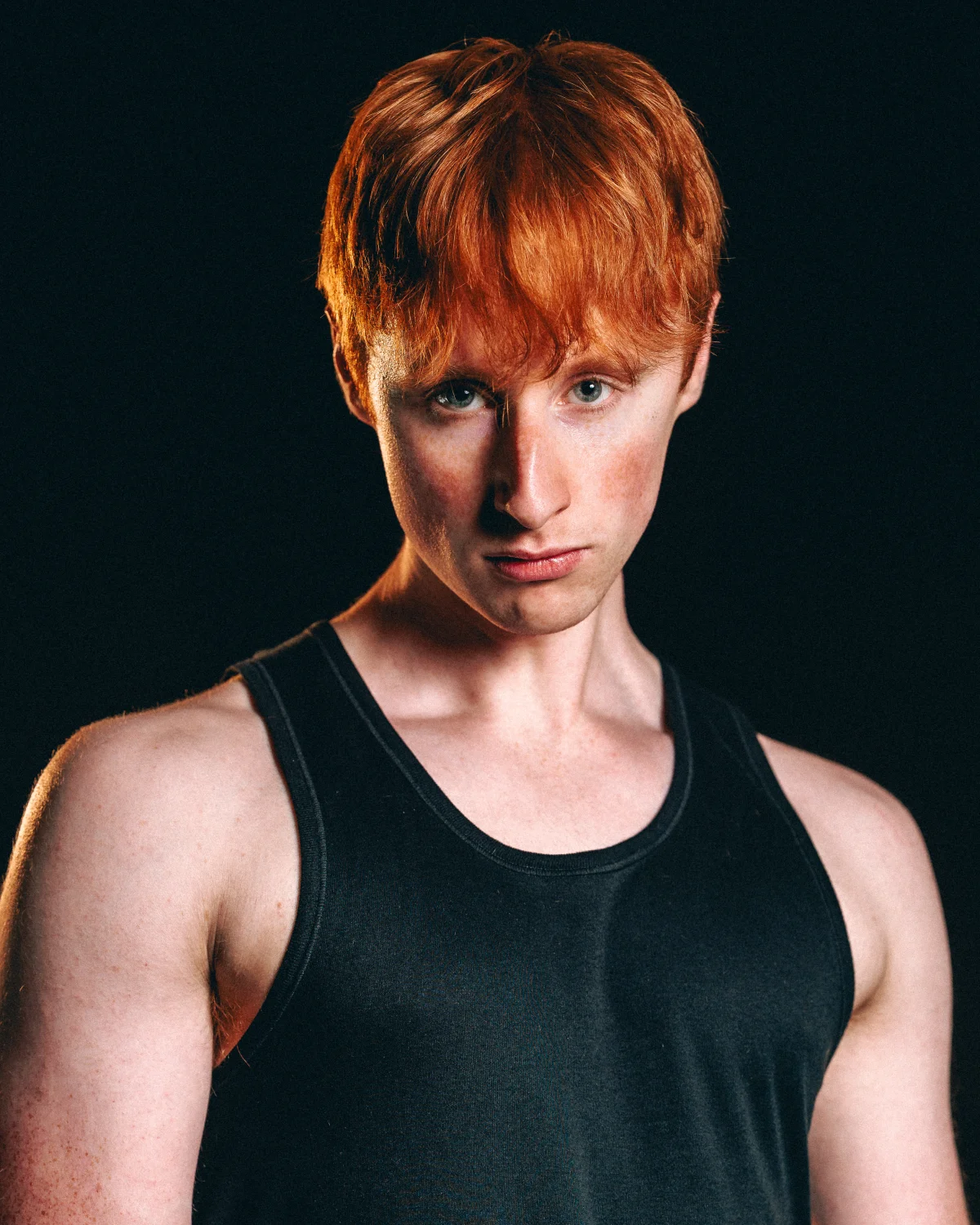 Close-up portrait of a young man with red hair in a black tank top against a dark background.