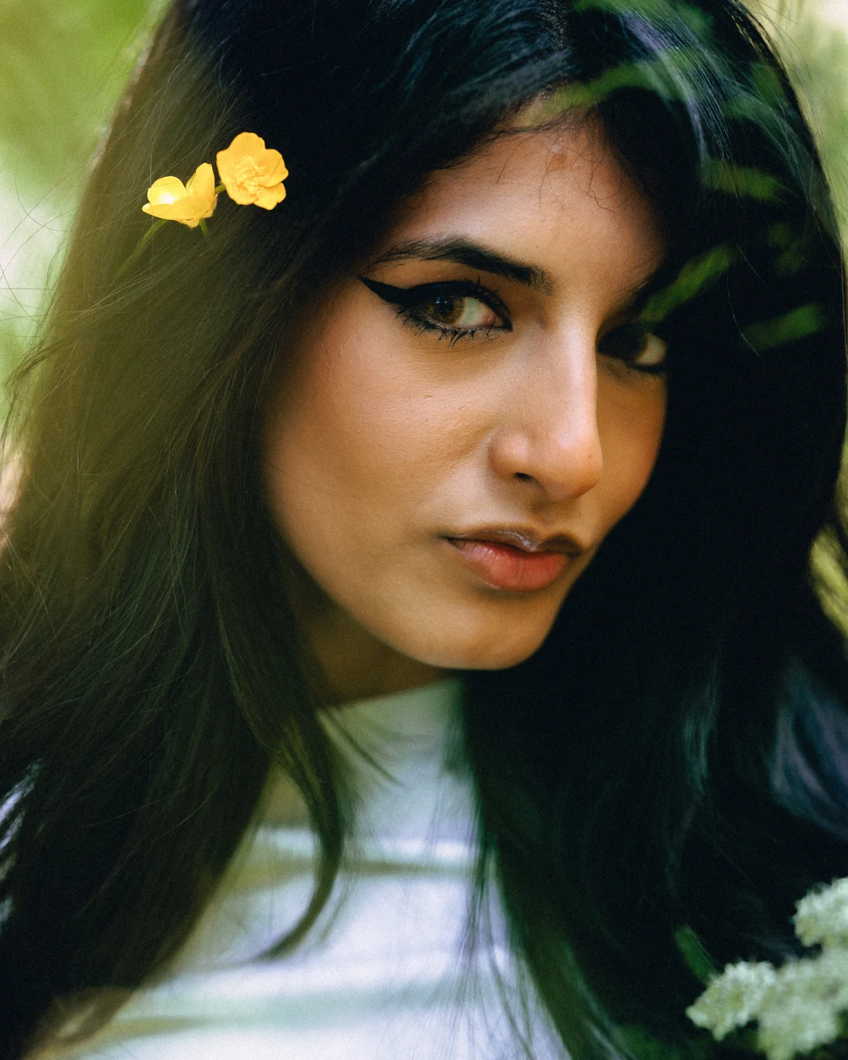 Close-up portrait of a woman with dark hair and yellow flowers in her hair, set against a soft green background.