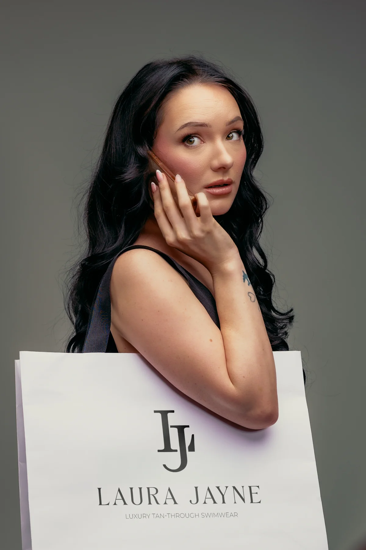 Woman with long dark hair posing with a phone behind a Laura Jayne luxury swimwear shopping bag.