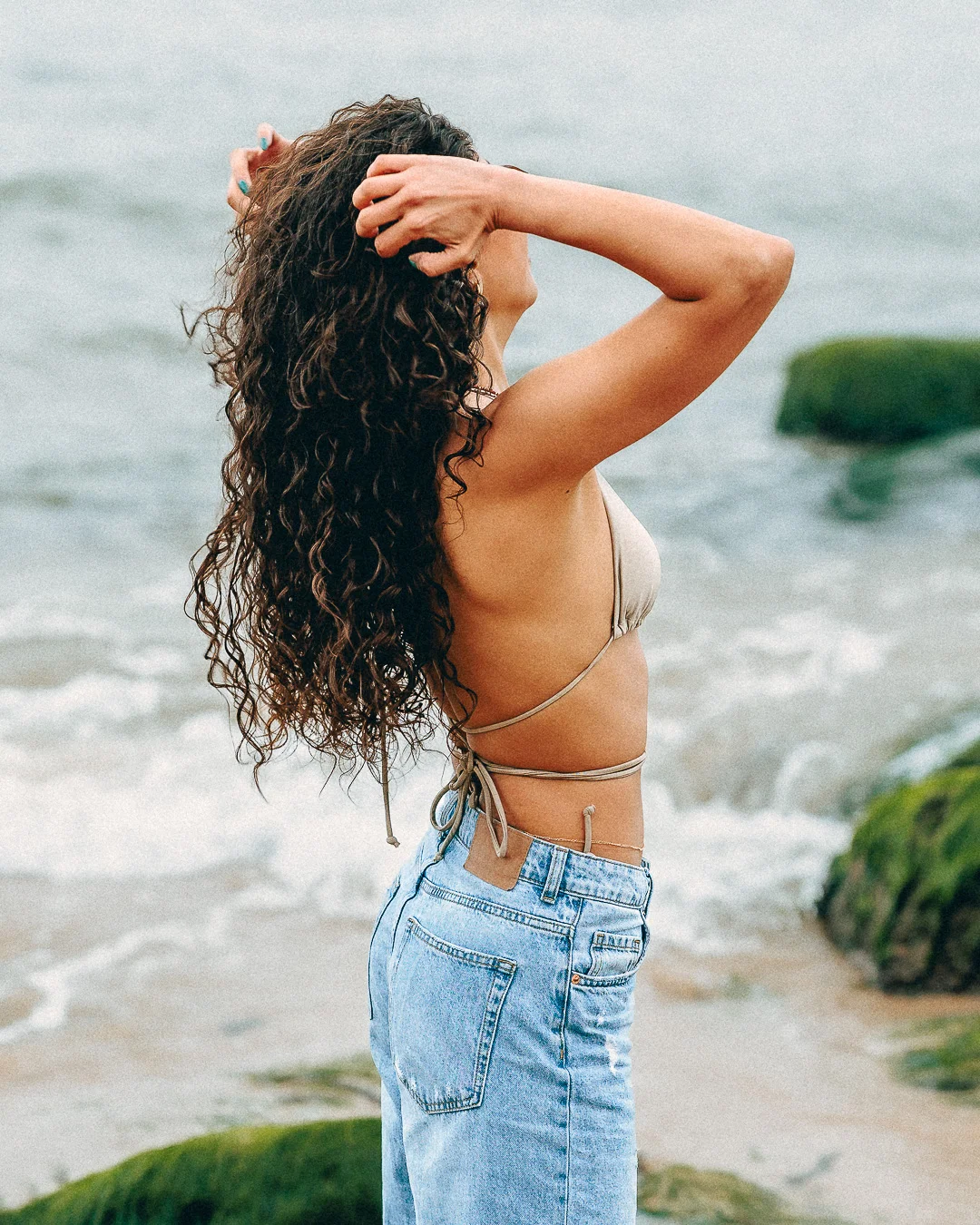 Woman with long curly hair posing on a beach wearing a bikini top and light blue jeans.