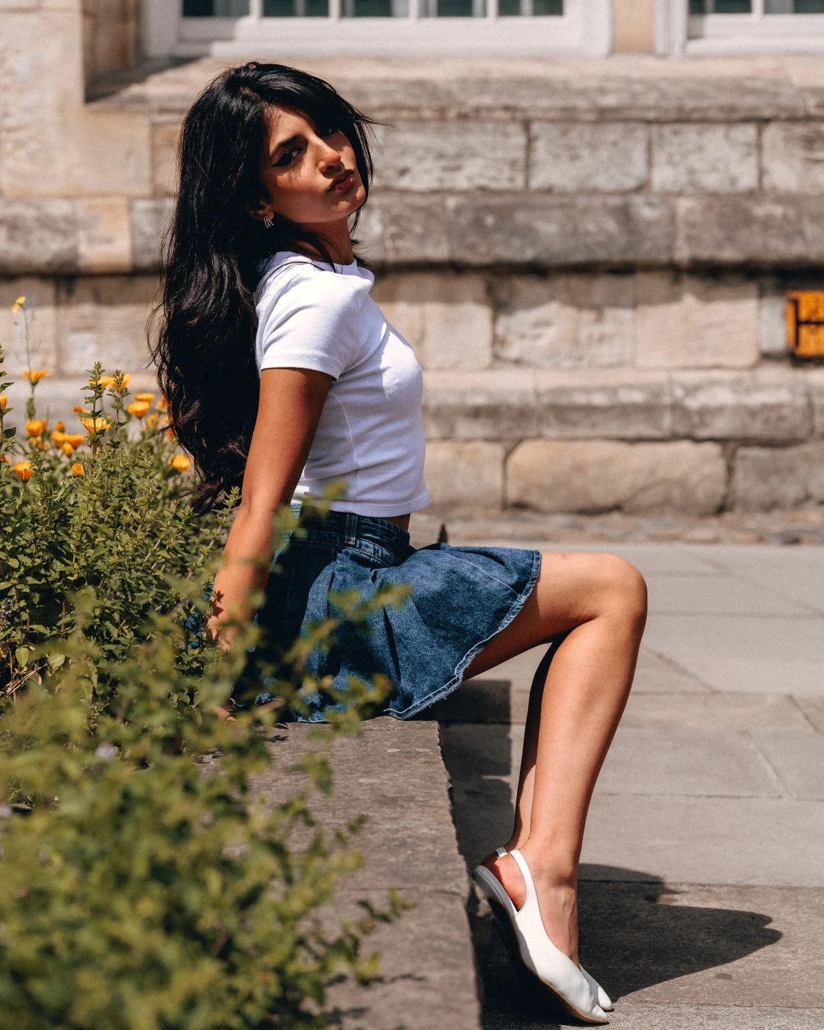 Young woman with long dark hair sitting on a stone ledge in a white t-shirt and denim skirt.