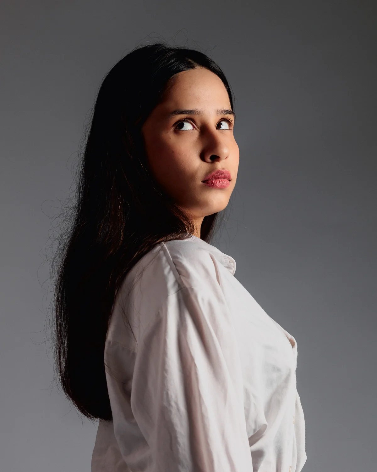 A young woman with long dark hair in a white shirt looking over her shoulder against a grey background.