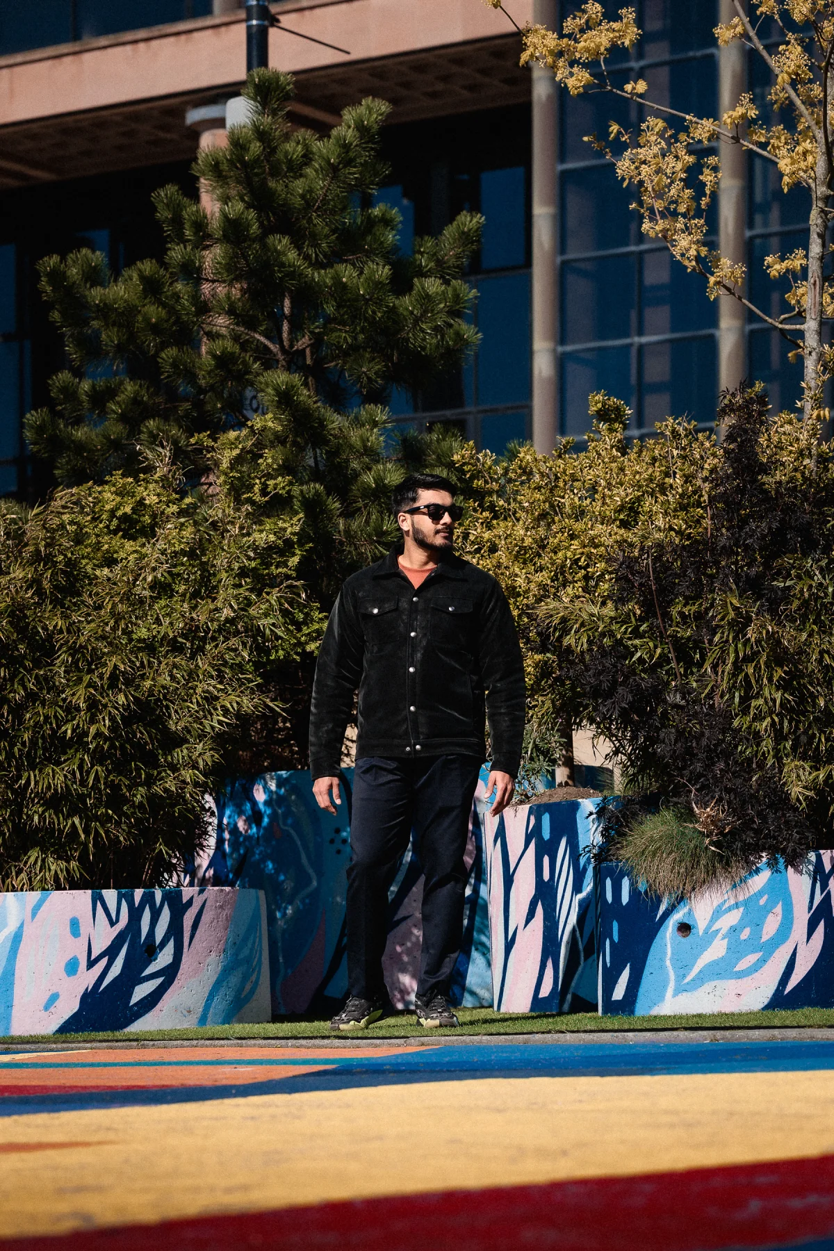 A man in sunglasses and a black jacket posing outdoors against greenery and a modern building.