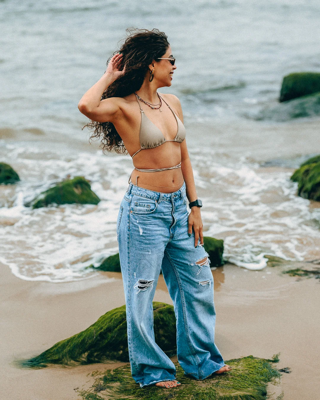 Woman with curly hair posing on a beach wearing a beige bikini top and ripped wide-leg jeans.