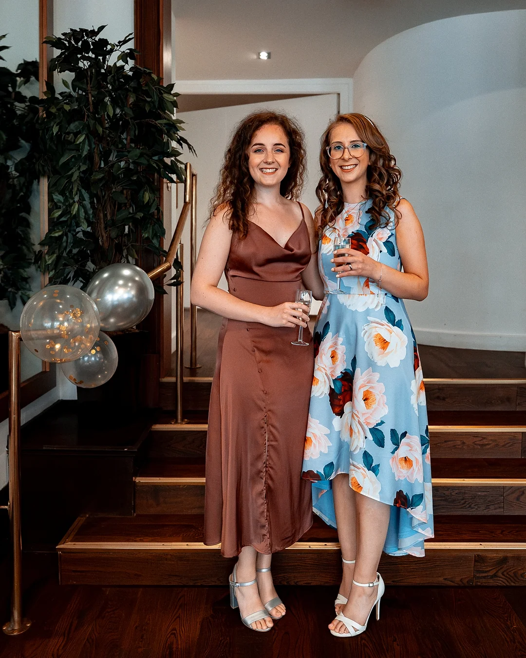 Two smiling women in elegant dresses holding champagne glasses, posing on a wooden staircase indoors.