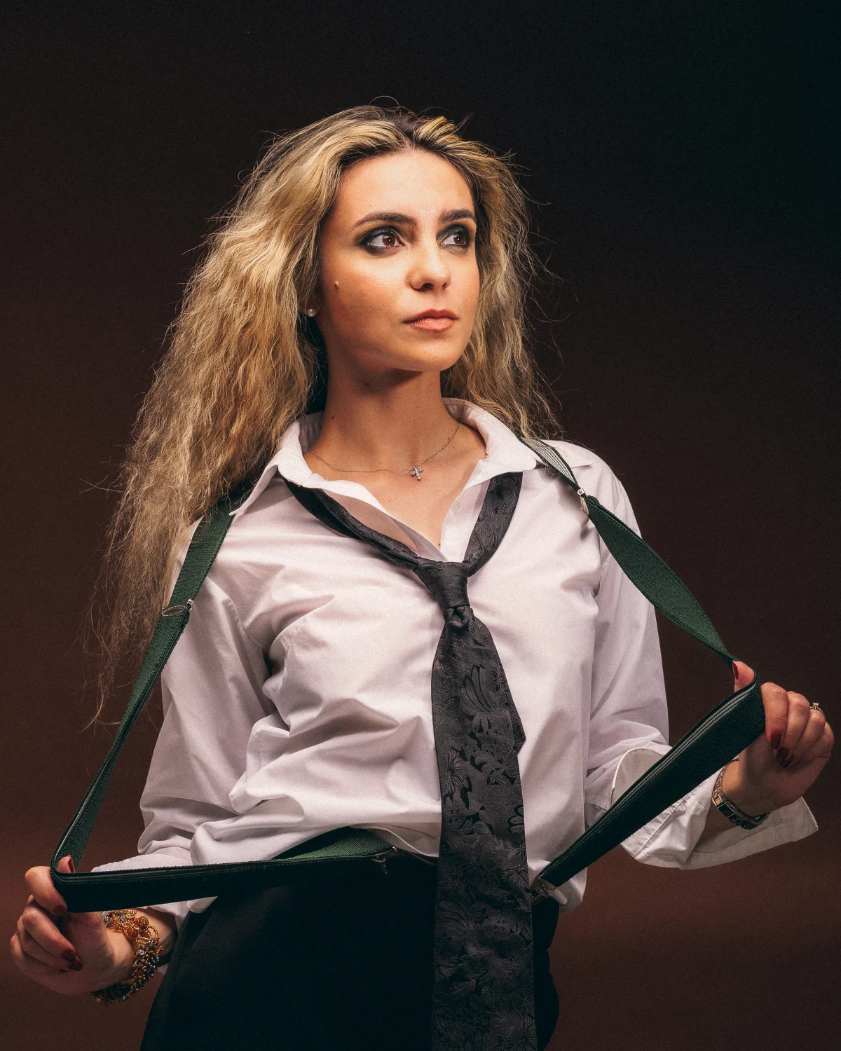 A woman with long blonde hair in a white shirt and loose tie, holding green suspenders against a dark background.