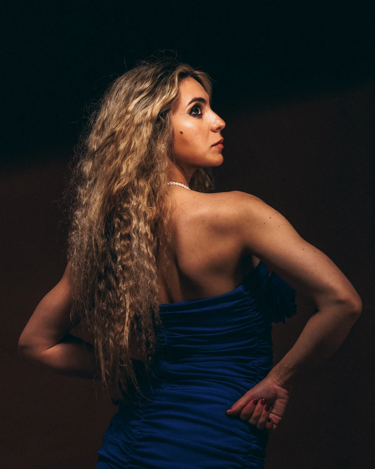 Side profile of a woman with long curly blonde hair in a blue strapless dress against a dark background.