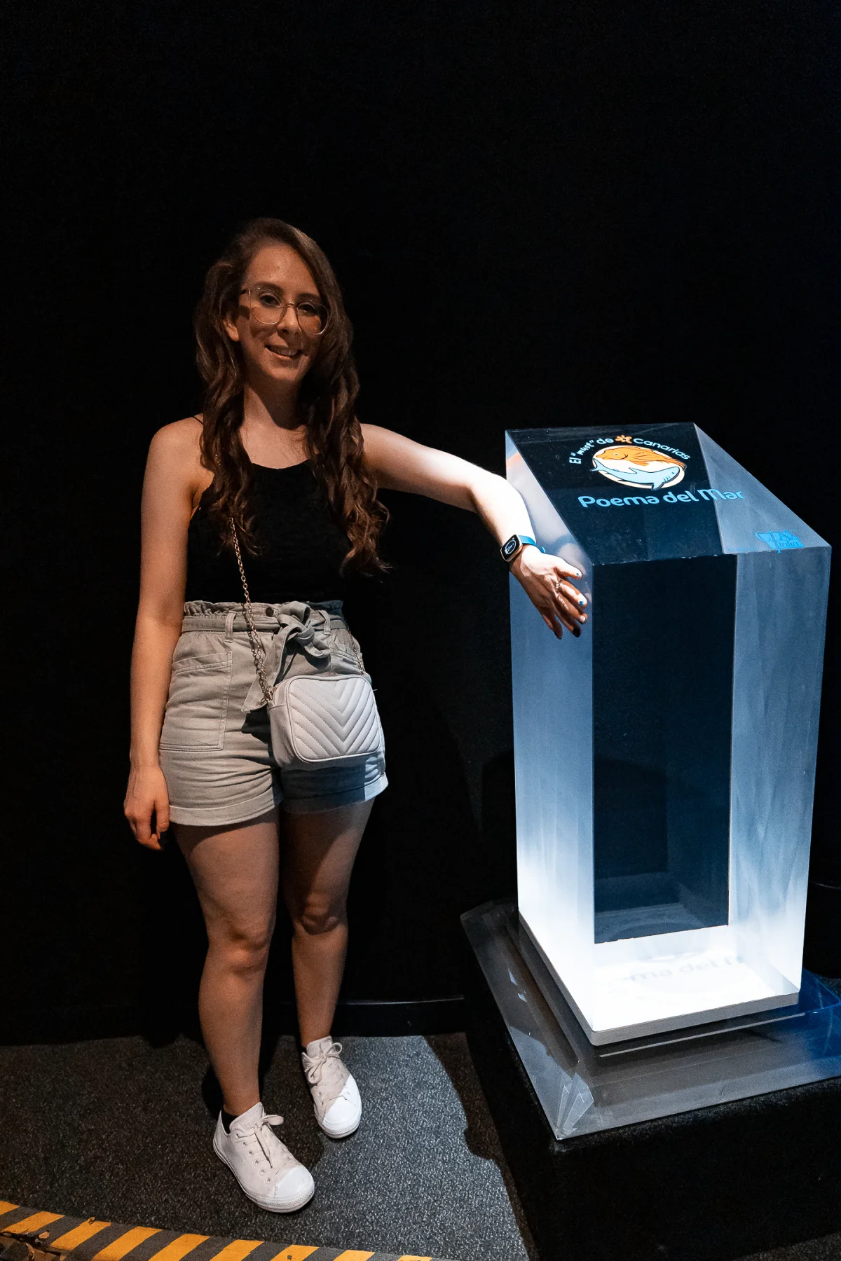 A smiling woman in a black top and grey shorts posing next to a glowing blue 'Poema del Mar' display case.