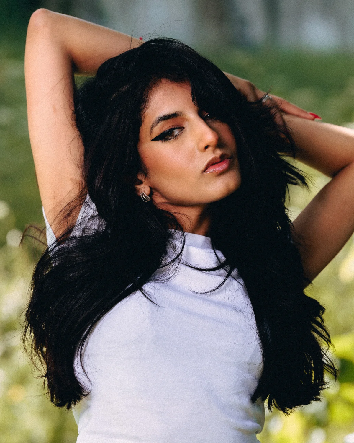 A young woman with long black hair and bold winged eyeliner posing outdoors in a white t-shirt.