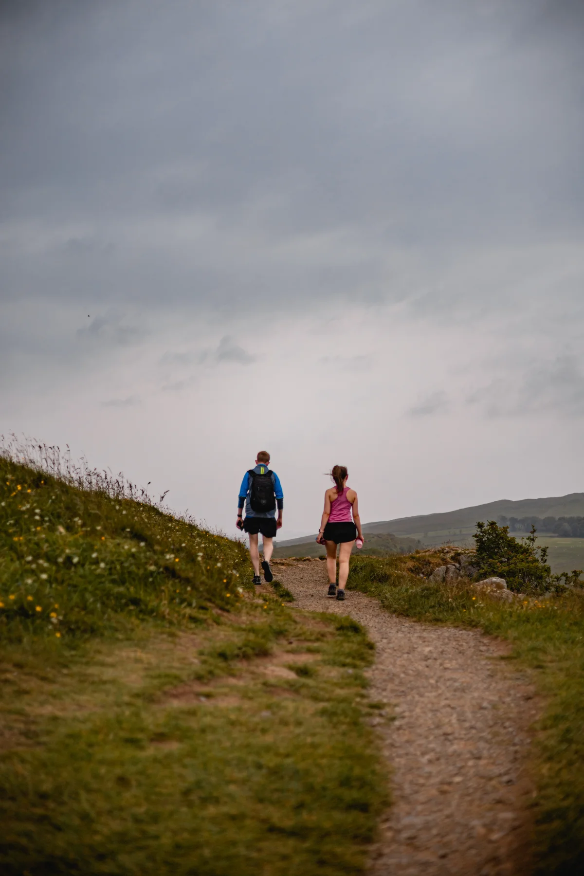 A man and woman walking away along a dirt path through green hills under an overcast sky.