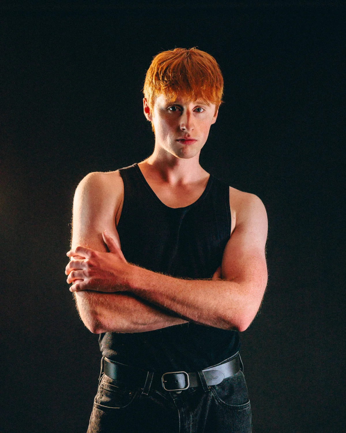 Studio portrait of a young man with red hair in a black tank top, arms crossed against a dark background.