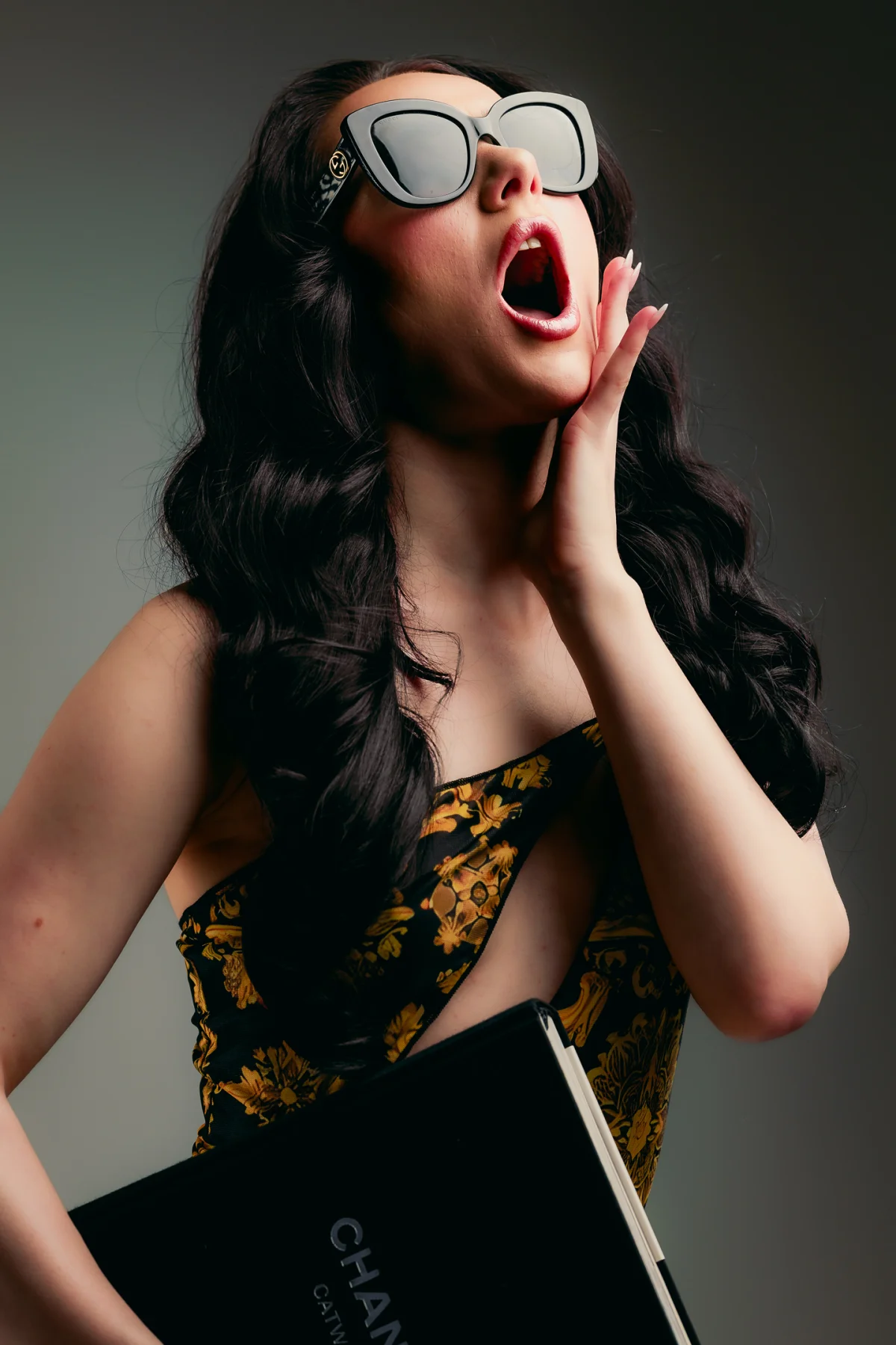 A woman in a yellow floral dress and oversized sunglasses looks surprised with her hand on her cheek.
