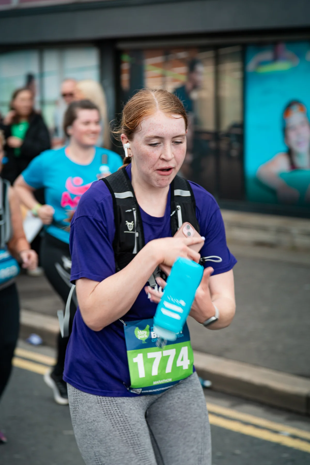 A woman in a purple t-shirt and running gear holding a blue water bottle during a marathon event.