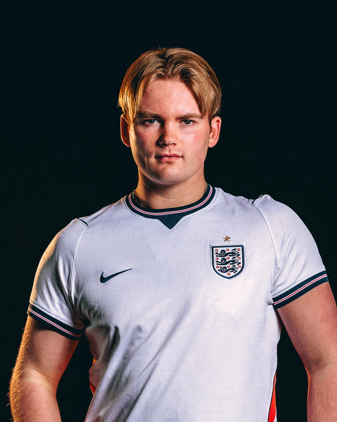 A young man with blond hair wearing an England football jersey against a black background.