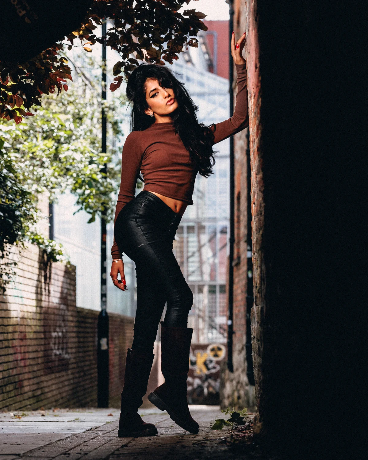 Woman in a brown crop top and black leather pants posing against an urban brick wall.
