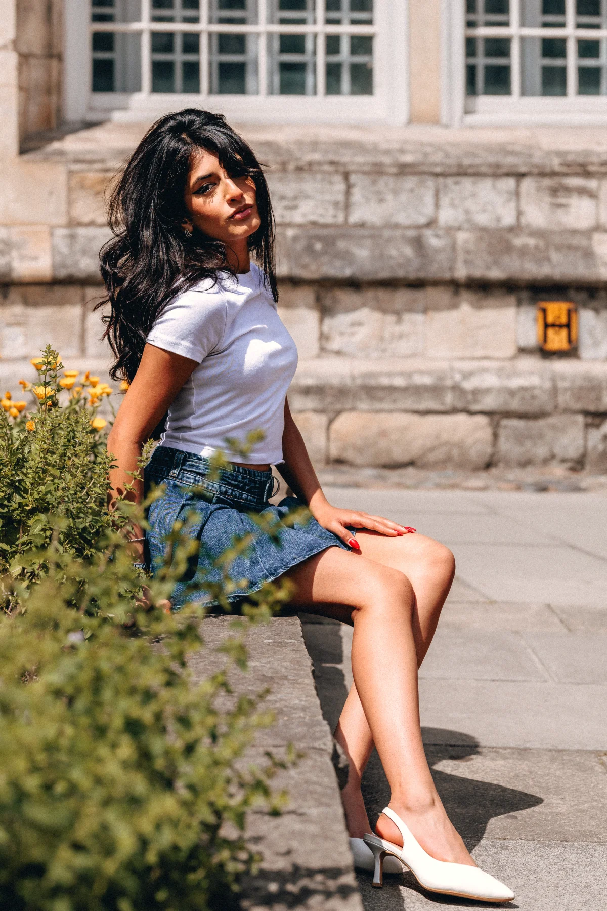 A young woman with long dark hair wearing a white t-shirt and denim mini skirt sitting on a stone ledge outdoors.