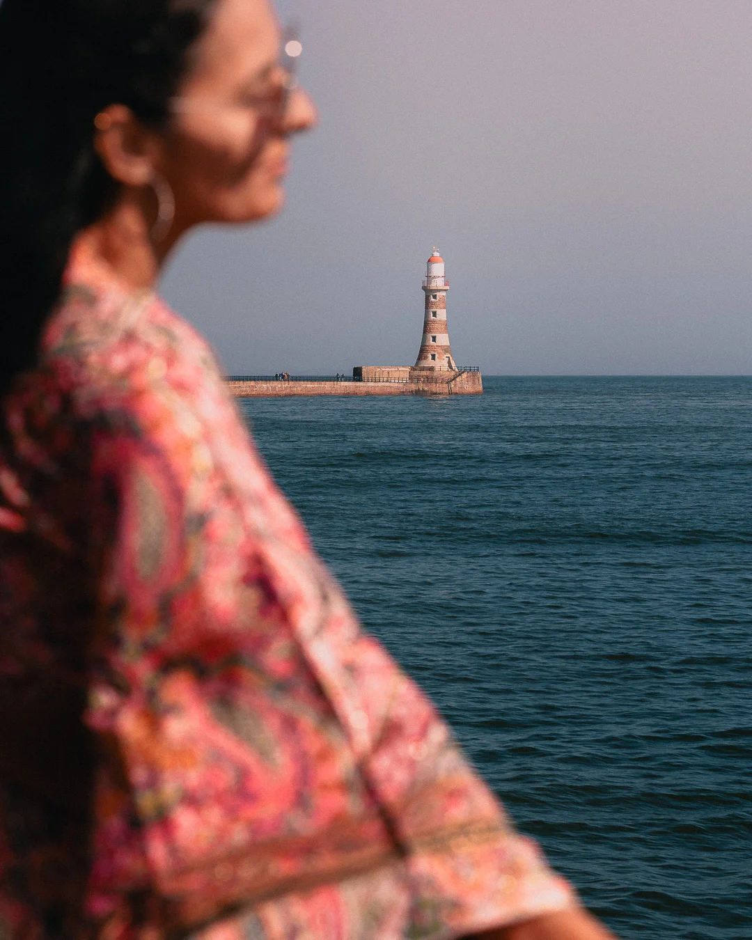Blurred profile of a woman in a colorful dress overlooking a distant lighthouse by the sea.