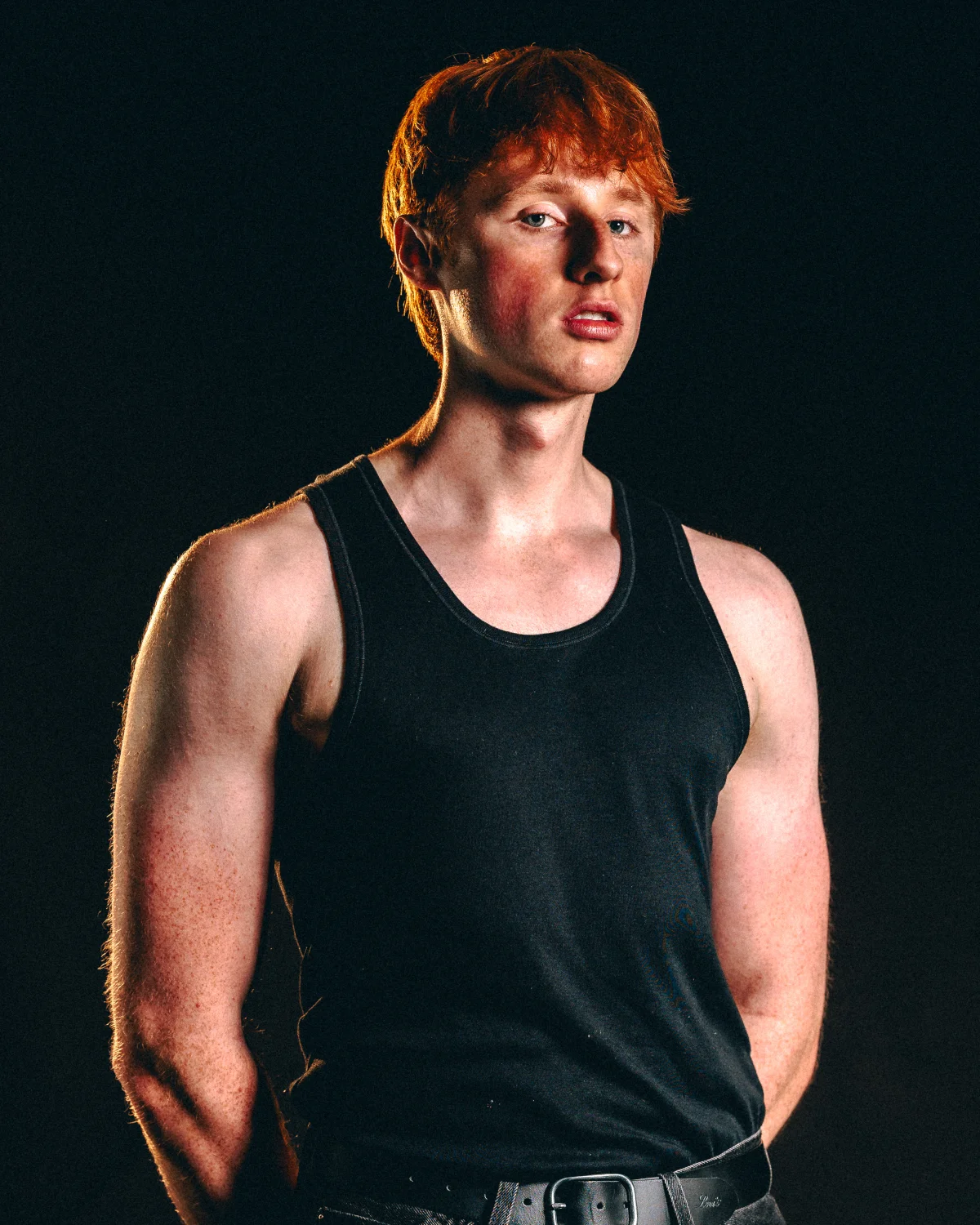 A young man with red hair in a black tank top posing against a dark background with moody studio lighting.