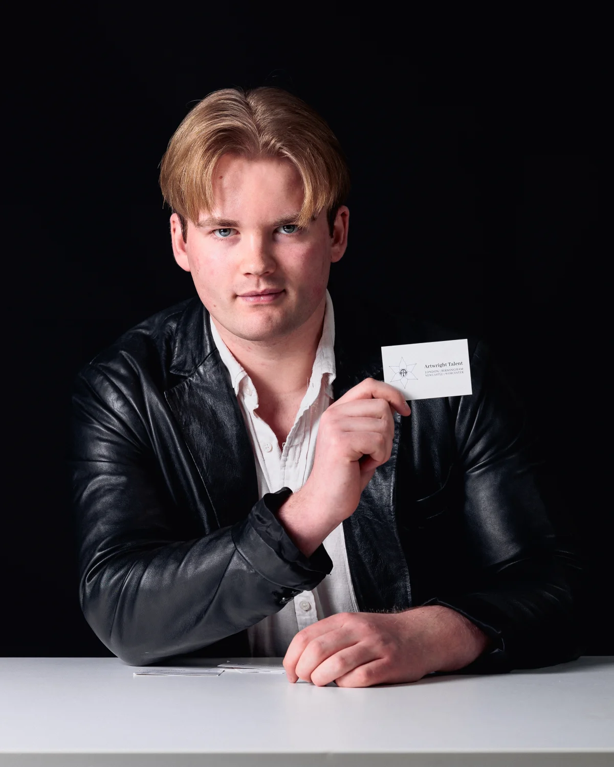 A young man with blond hair in a black leather jacket holding a business card against a dark background.