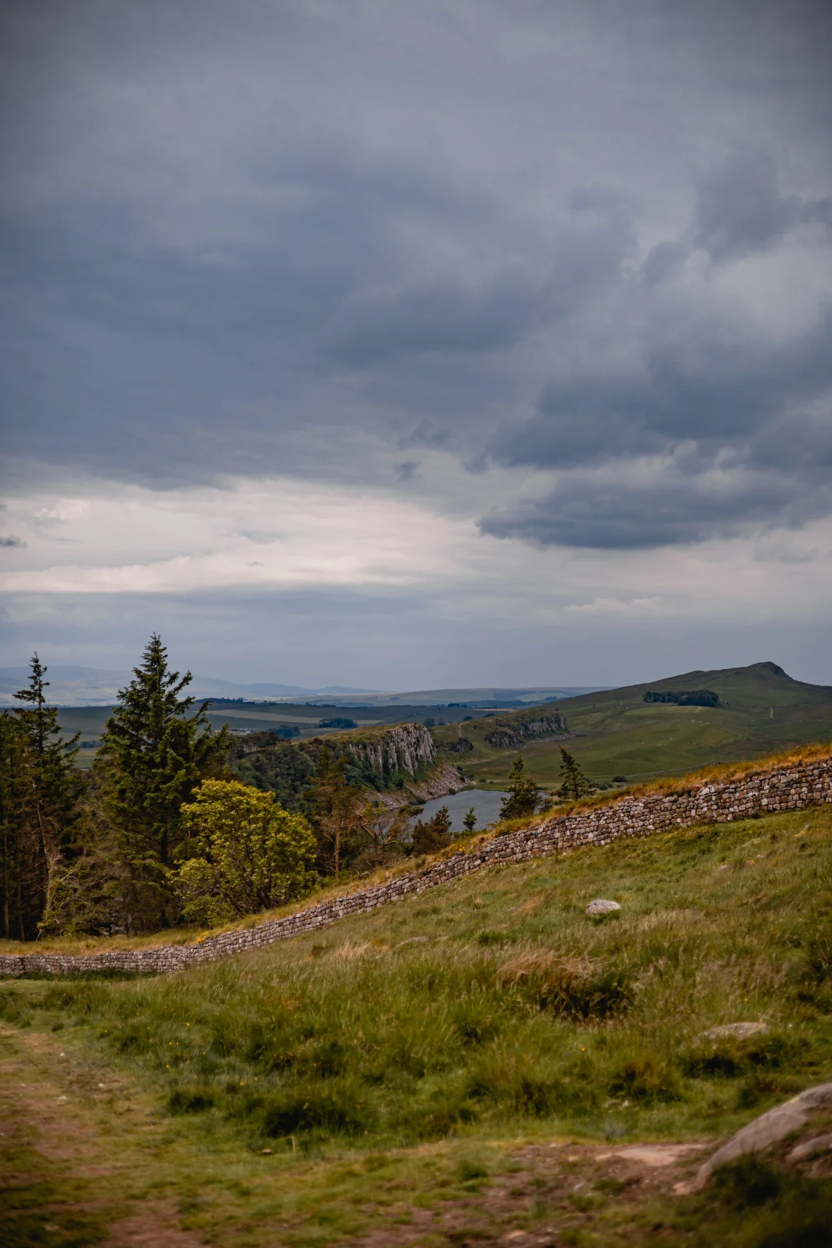 A moody landscape with rolling green hills, a dry stone wall, and evergreen trees under a dramatic overcast sky.