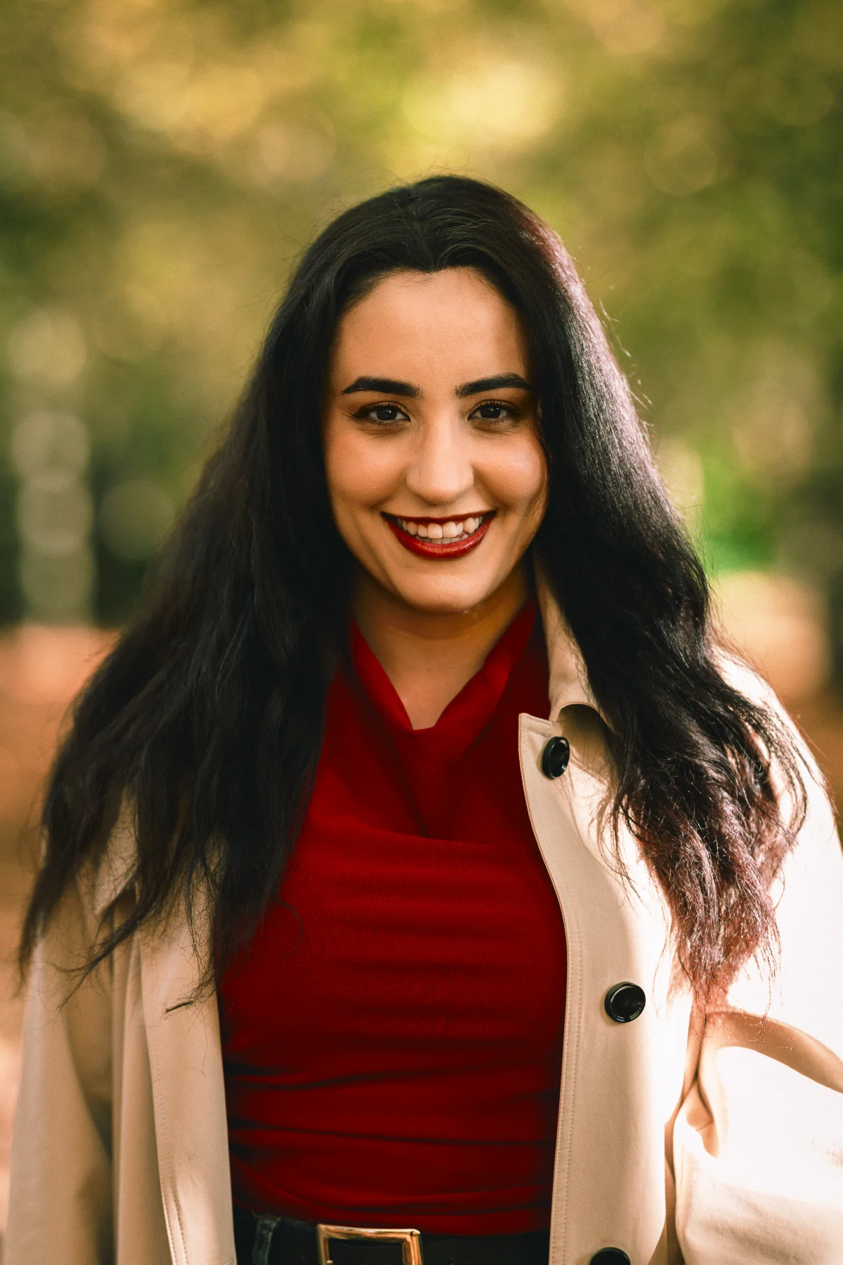 A smiling woman with long dark hair wearing a red top and beige coat in an outdoor autumn setting.