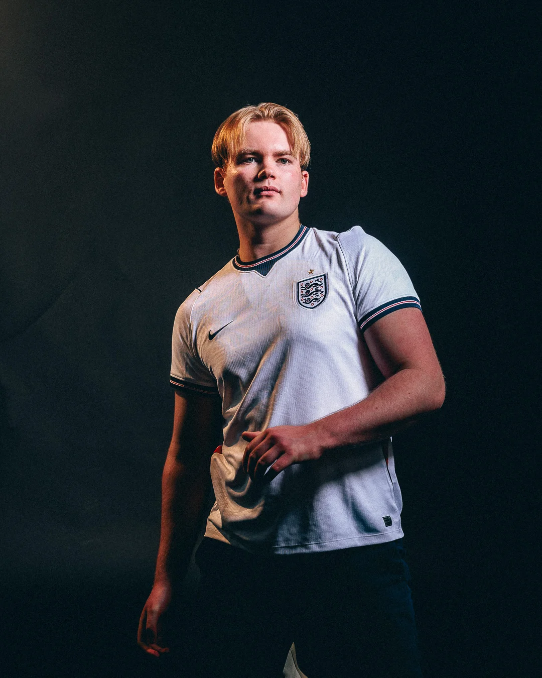 A young man with blond hair wearing an England football jersey posing against a dark studio background.