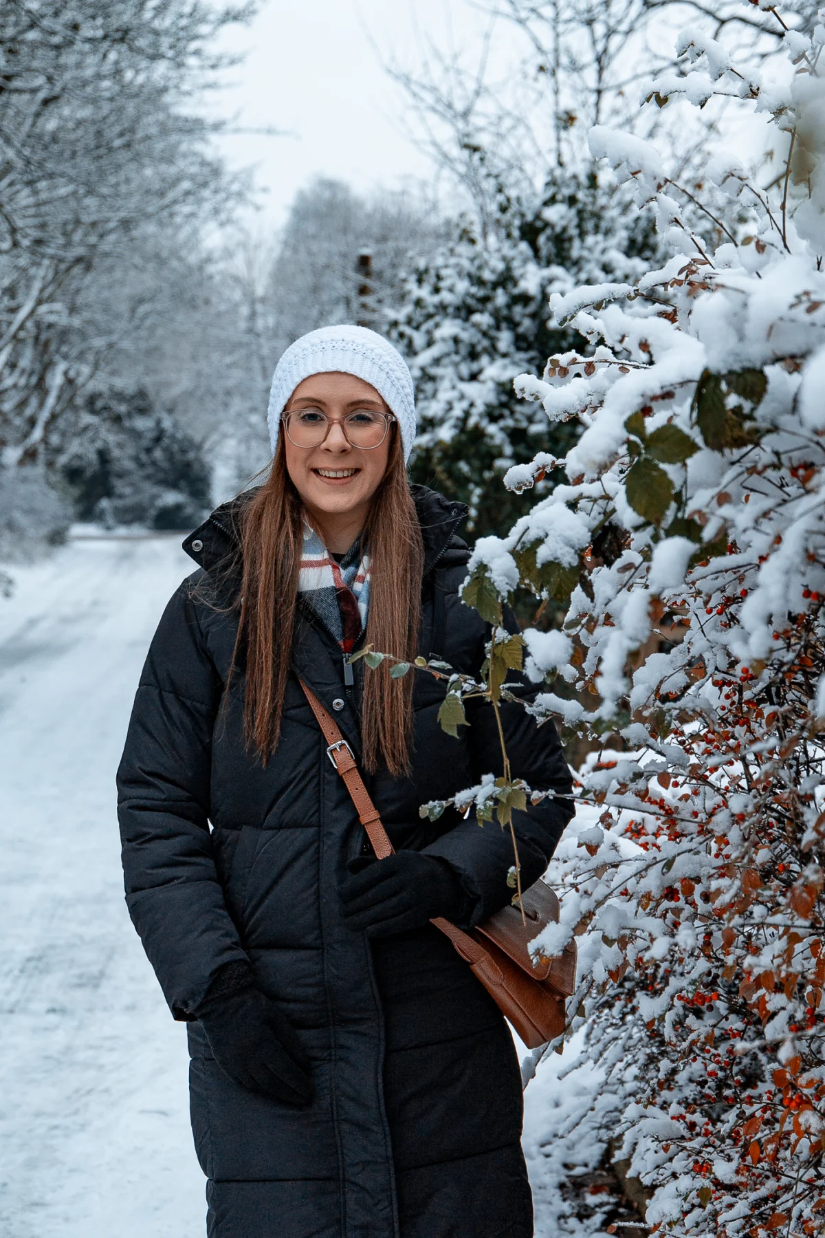 A smiling woman in a white beanie and black winter coat standing amidst snow-covered bushes.