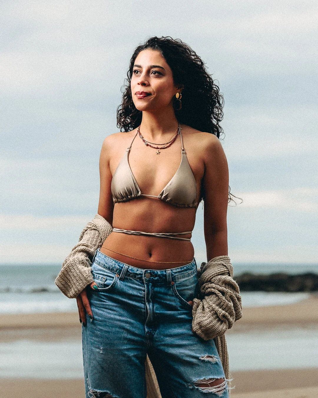 Woman with curly hair posing on a beach wearing a gold bikini top and blue jeans.