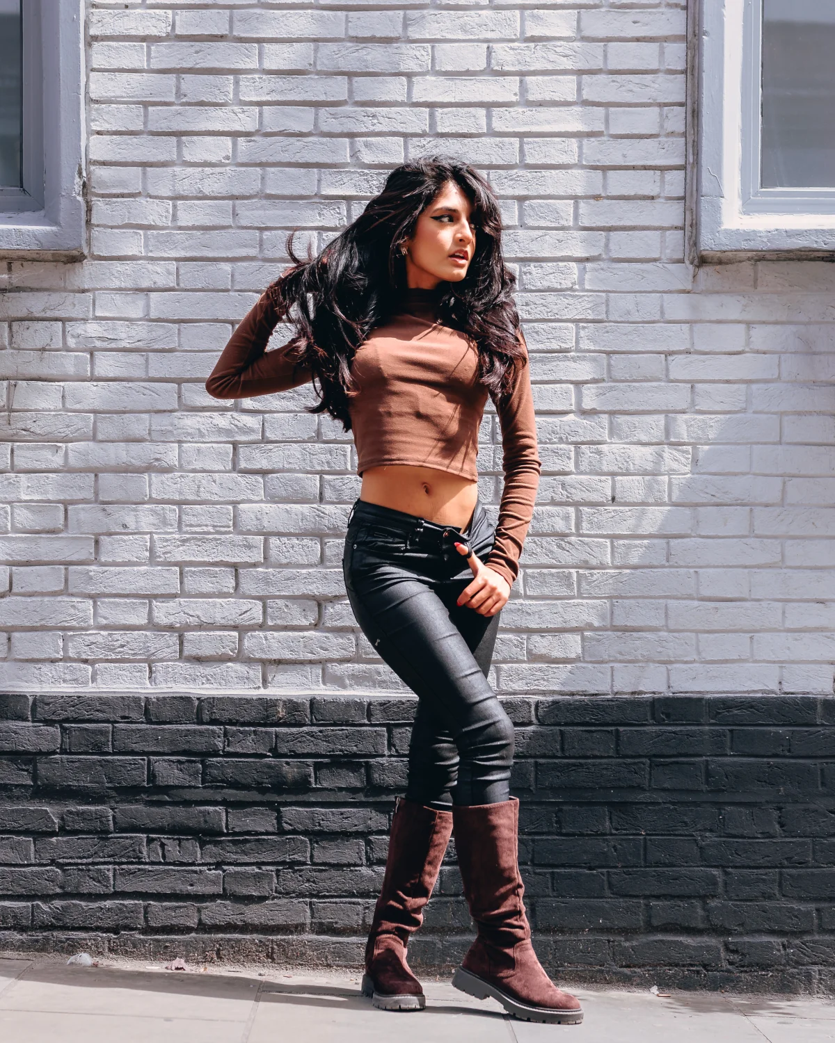 A woman with long dark hair in a brown crop top and black leather pants posing against a white brick wall.
