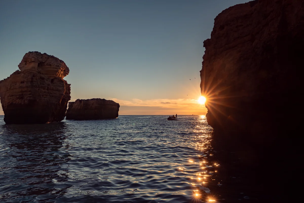 A serene sunset scene featuring dramatic coastal rock formations and a small boat on calm waters.