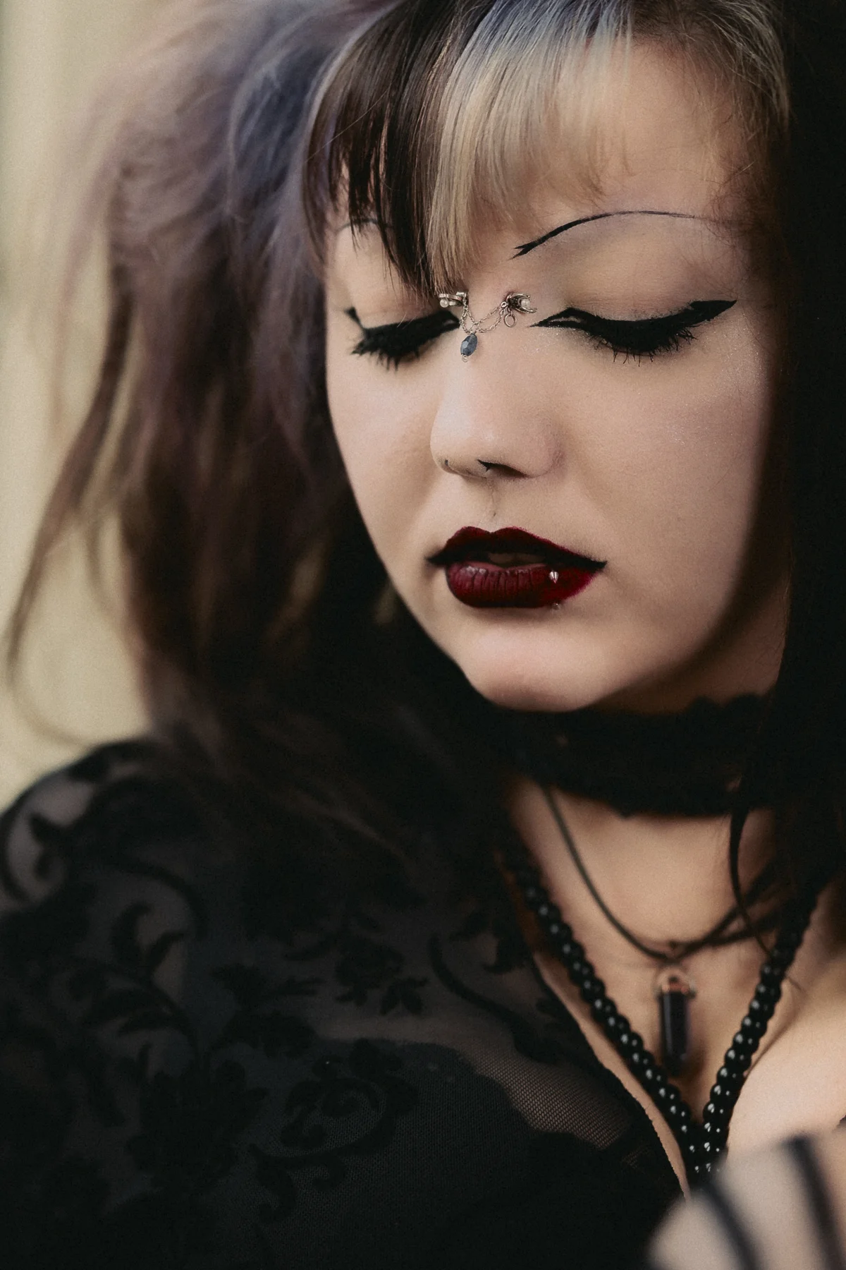 Close-up portrait of a person with gothic makeup, dark red lipstick, and face jewelry, eyes closed.