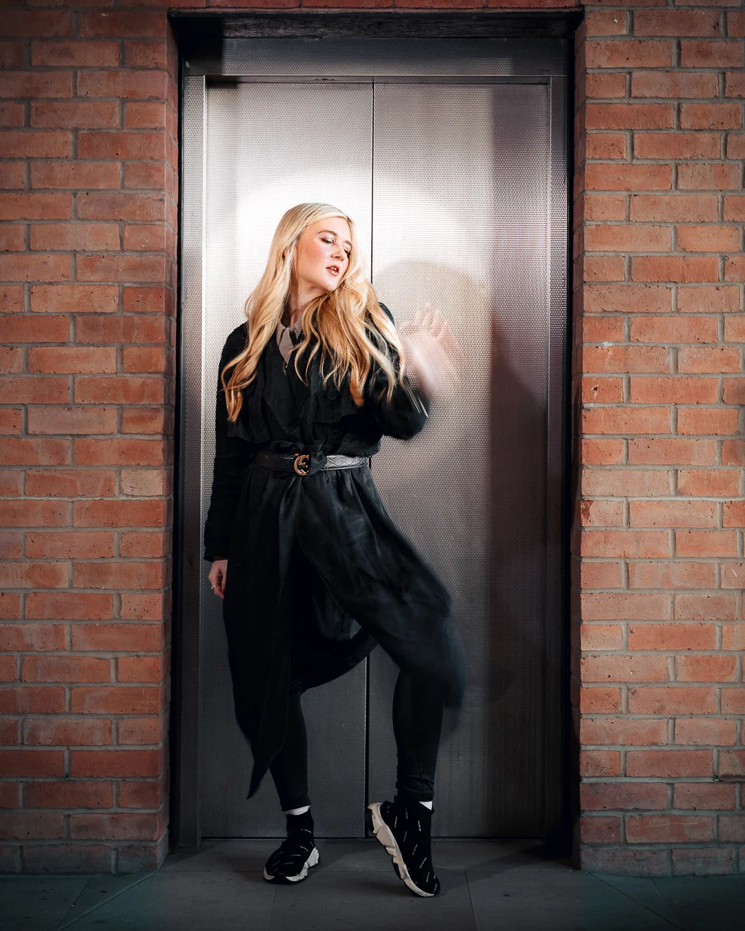 Woman with long blonde hair in a black belted dress and sneakers posing before elevator doors and brick walls.