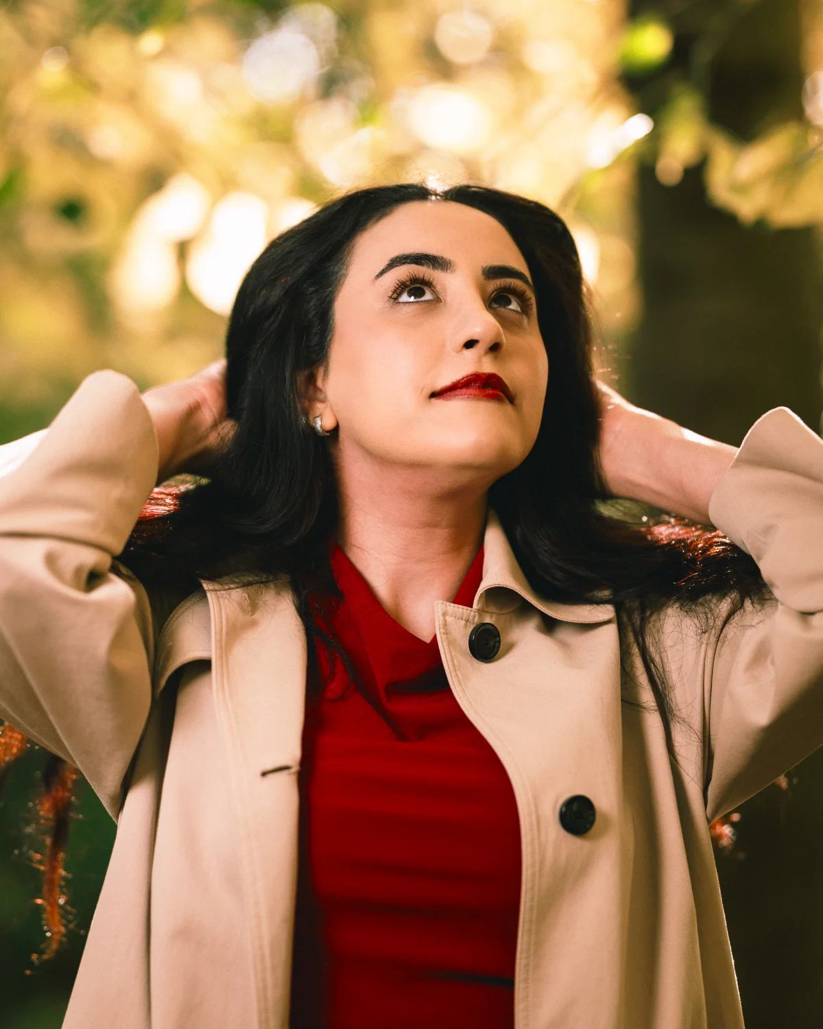 Woman with dark hair and red lipstick in a beige trench coat looking up against a blurred outdoor background.