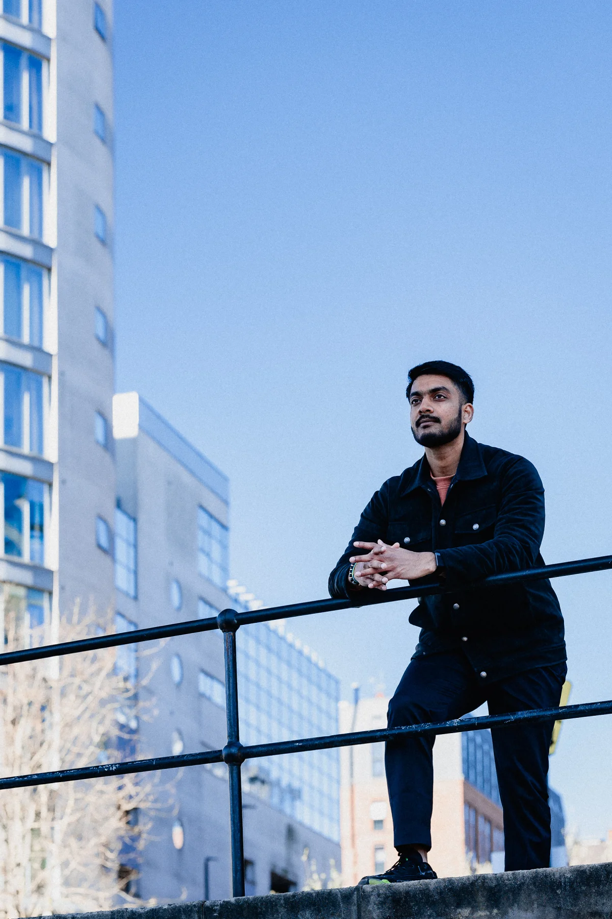 A young man with a beard wearing a black jacket leaning on a metal railing in an urban setting under a clear blue sky.