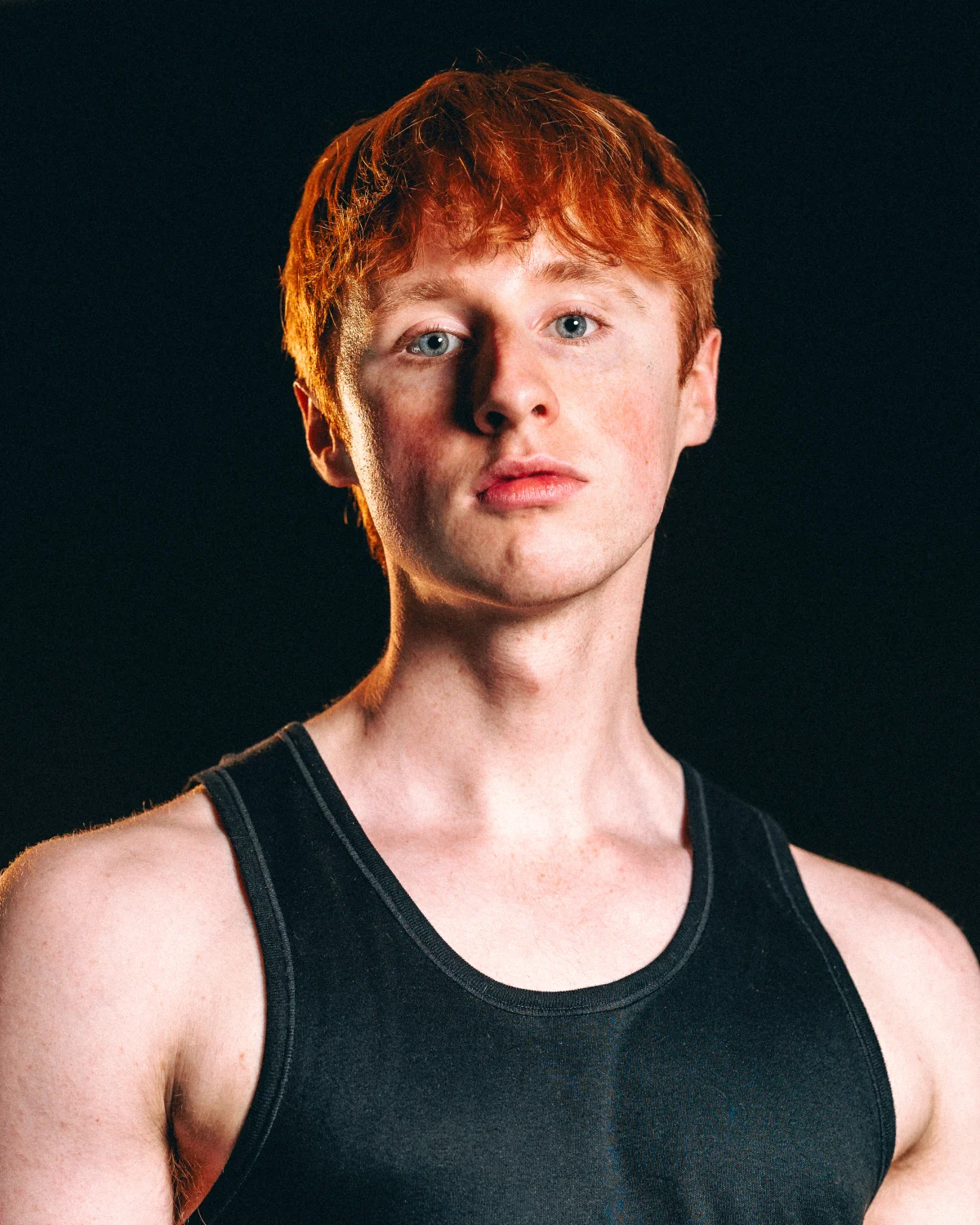 Close-up portrait of a young man with red hair and blue eyes wearing a black tank top against a dark background.